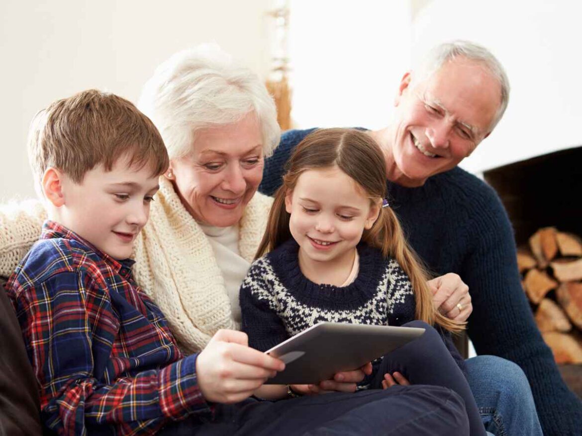 Avós e duas crianças reunidos no sofá olhando um tablet  com fotos