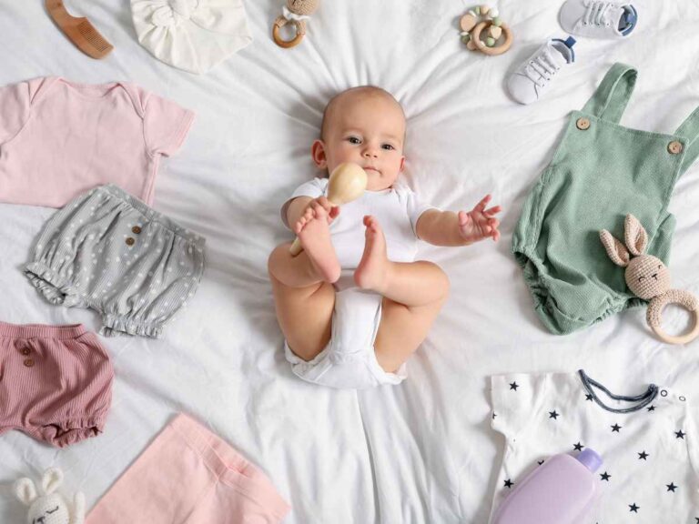 Bebê deitada sobre cama branca cercado por peças de roupa, acessórios e itens do enxoval
