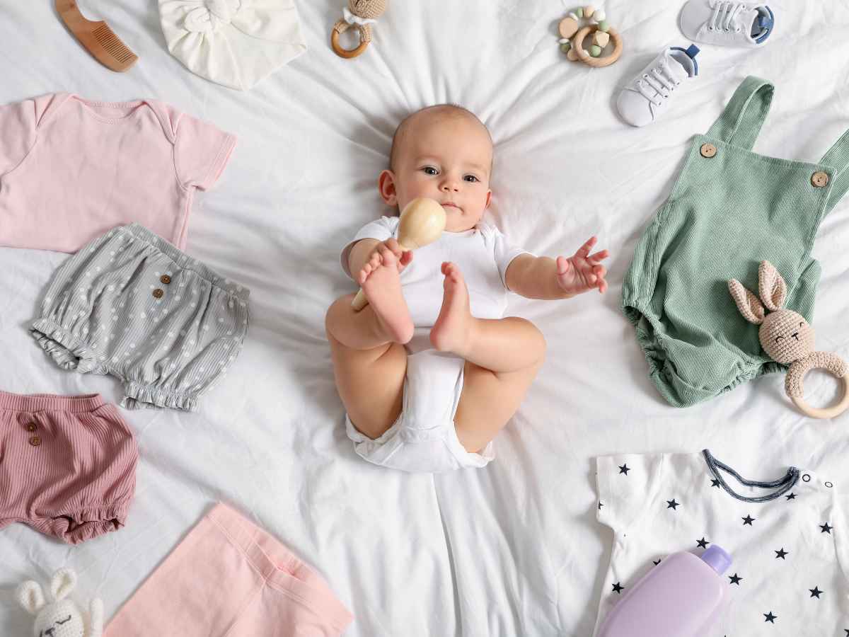 Bebê deitada sobre cama branca cercado por peças de roupa, acessórios e itens do enxoval