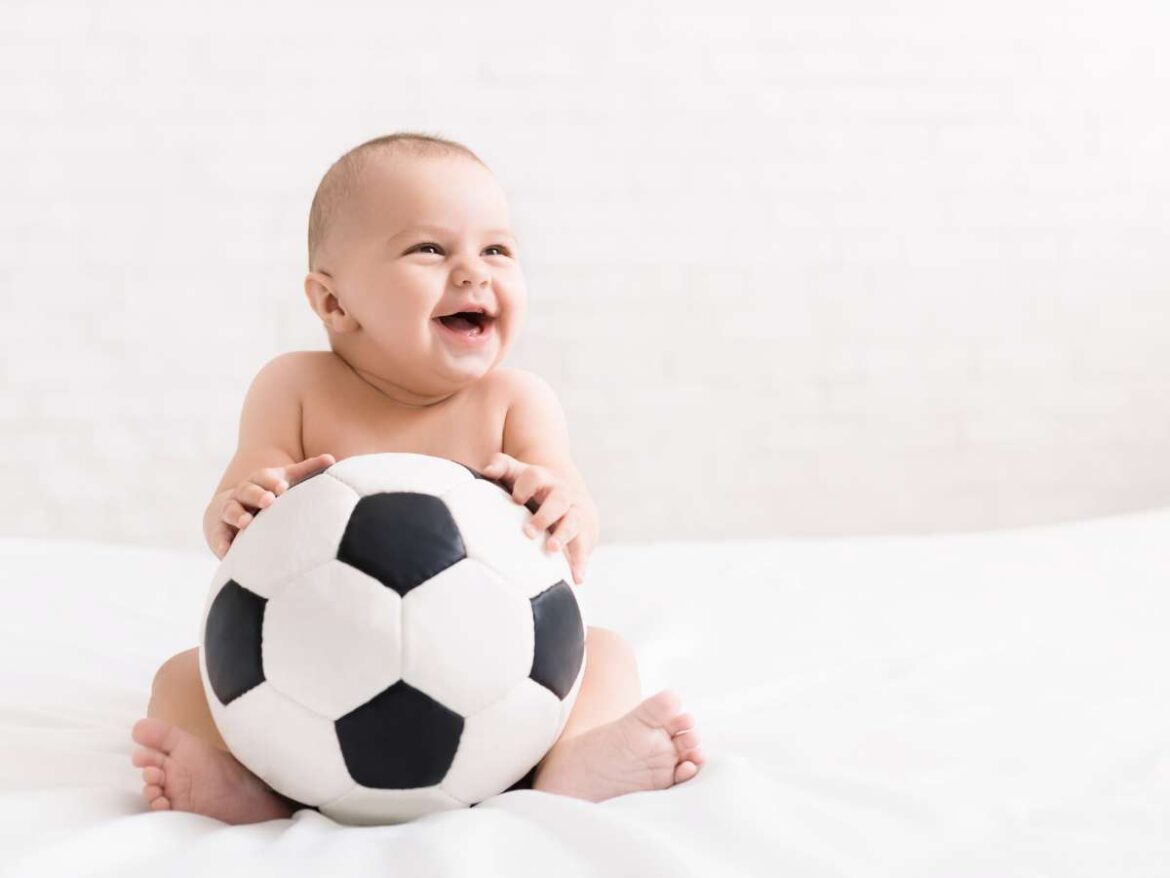 Bebê sorridente sentado segurando uma bola de futebol em fundo claro.