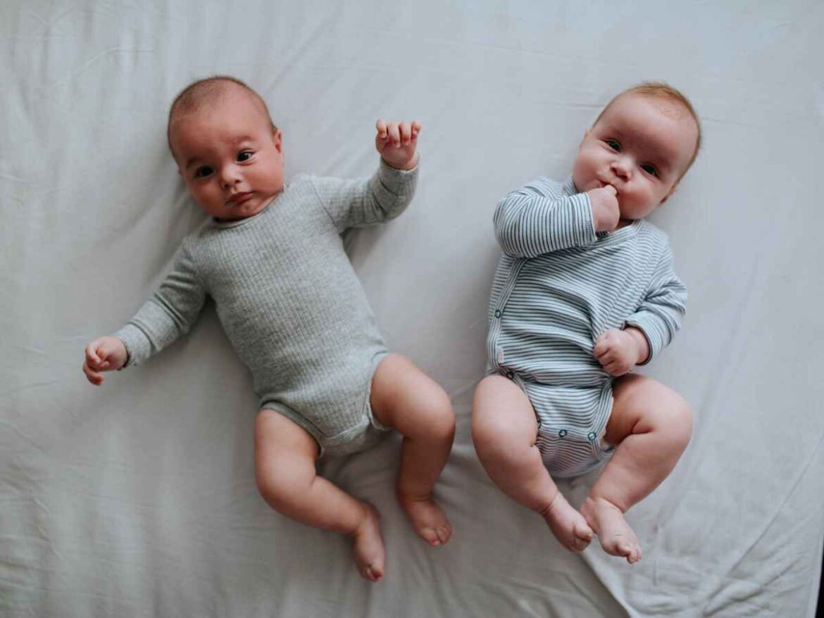 Dois bebês gêmeos deitados sobre tecido claro usando bodies confortáveis em tons neutros
