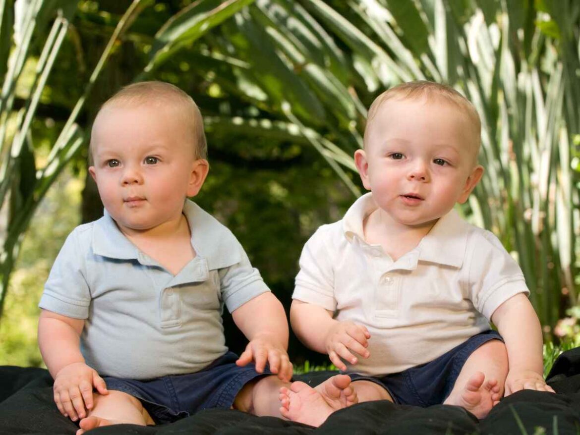 Bebês gêmeos sentados em área externa com roupas casuais em tons claros