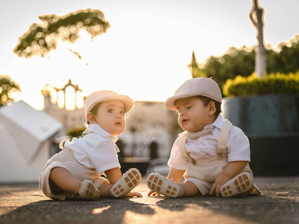 Bebês gêmeos sentados ao ar livre usando roupas sociais claras e bonés