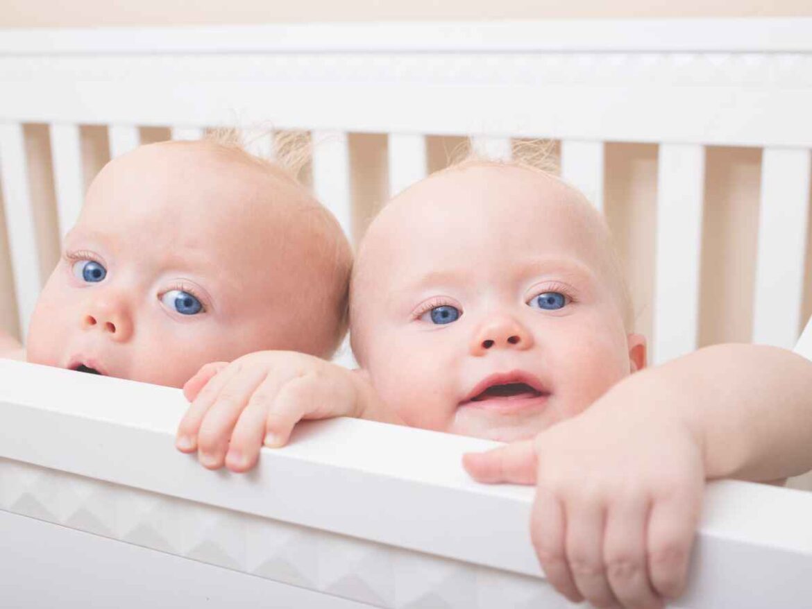 Dois bebês gêmeos apoiados na grade do berço olhando para frente