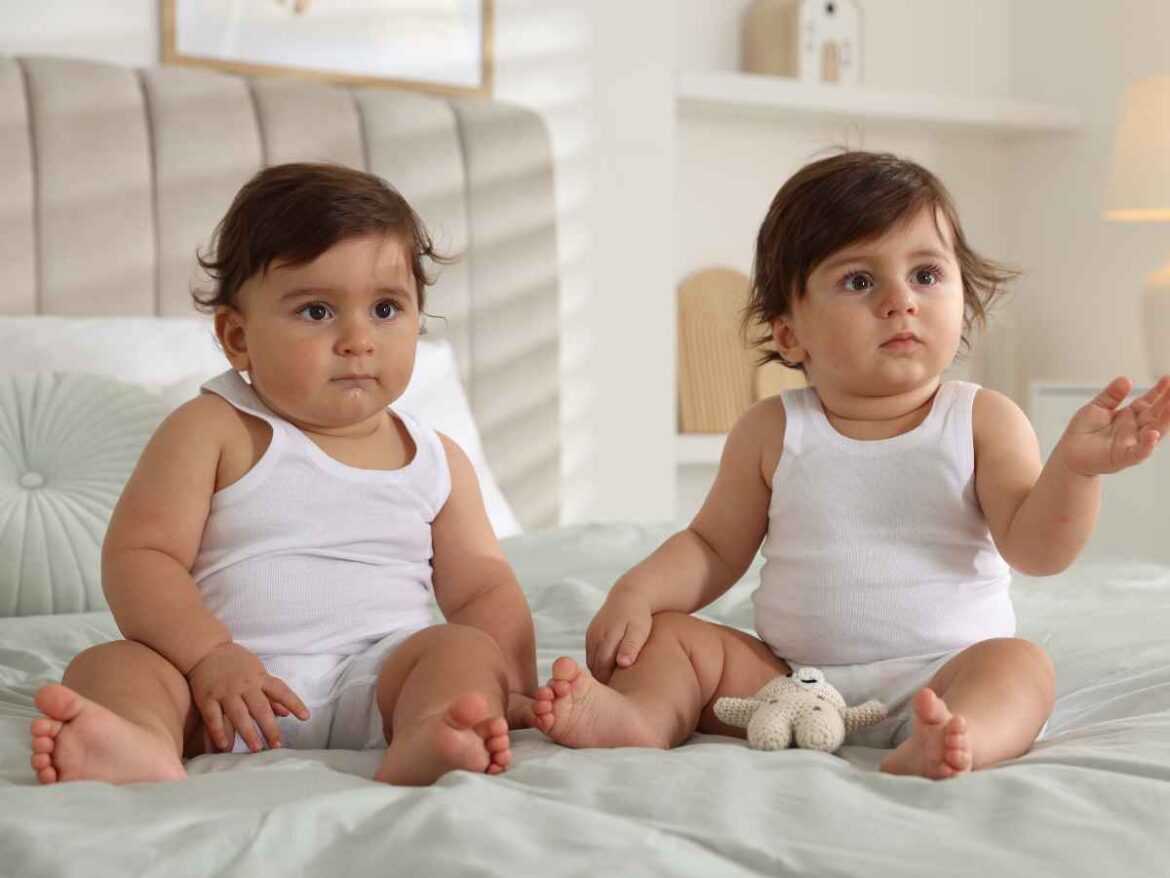 Dois bebês gêmeos sentados sobre a cama usando roupas básicas brancas