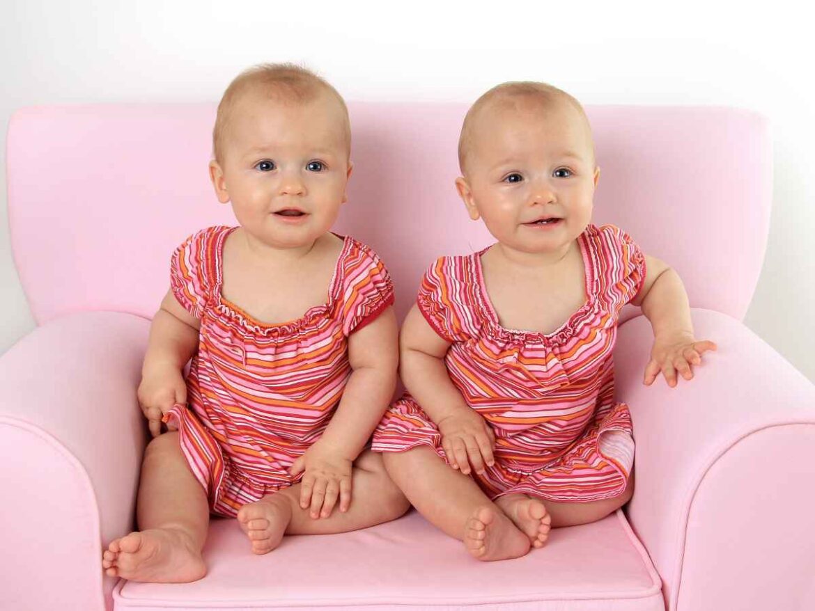 Bebês gêmeas sentadas em poltrona rosa usando vestidos listrados iguais