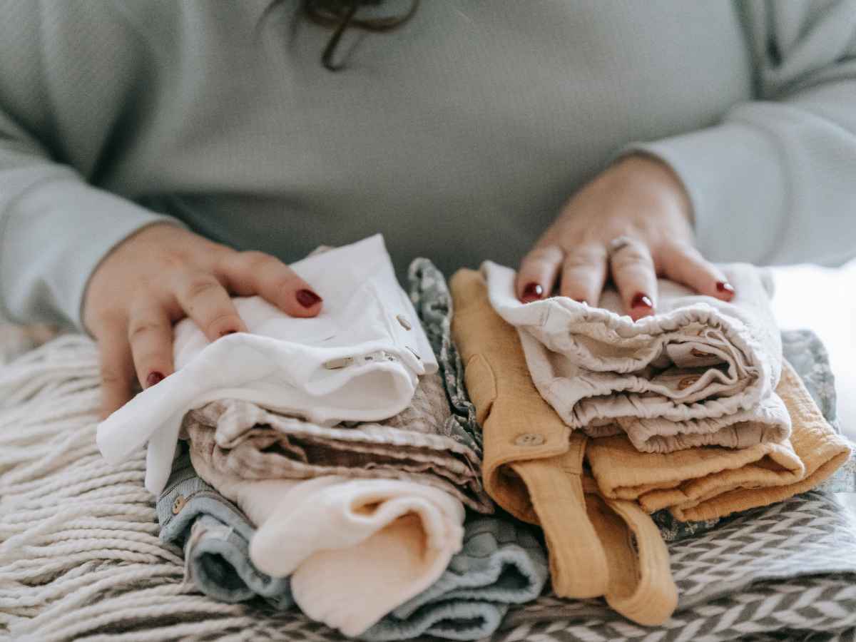 Pessoa organizando e dobrando roupas de bebê em tons neutros sobre a cama