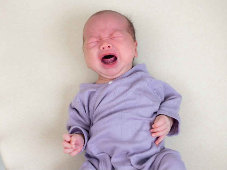 Bebê recém-nascido vestido com roupa lilás, deitado sobre uma superfície clara, chorando com os olhos fechados e expressão de desconforto