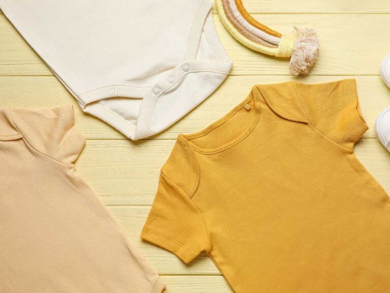 Roupas de bebê em tons amarelo e bege posicionados contra fundo de madeira.