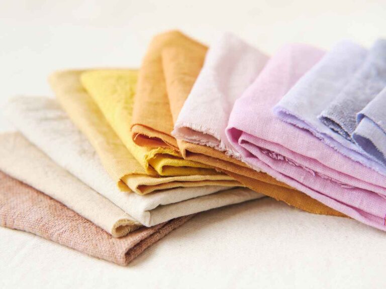 Conjunto de tecidos dobrados em tons suaves de bege, amarelo, rosa e lilás, organizados sobre uma superfície clara, destacando diferentes texturas e opções de materiais