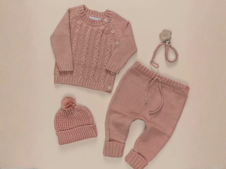 Conjunto de peças de tricô para bebê em tom rosa, composto por blusa de manga longa, calça e touca com pompom, acompanhado de prendedor de chupeta.
