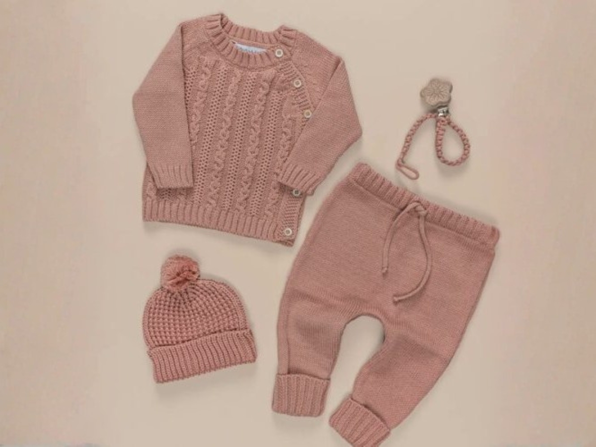 Conjunto de peças de tricô para bebê em tom rosa, composto por blusa de manga longa, calça e touca com pompom, acompanhado de prendedor de chupeta.
